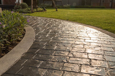 close up picture of a long stamped concrete walkway in three colors bordering a lush green area.